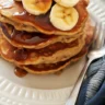 Fluffy whole wheat banana pancakes stacked on a plate with maple syrup and fresh bananas
